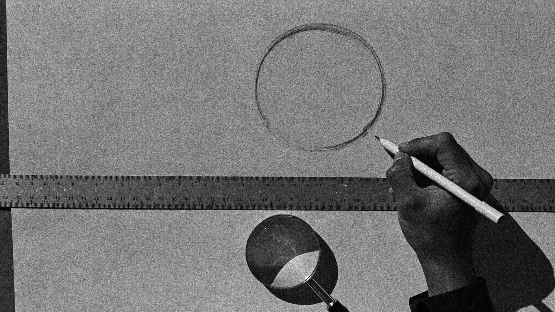 A hand drawing a careful circle in pencil beside a steel ruler and a loupe, the page being corrected by hand