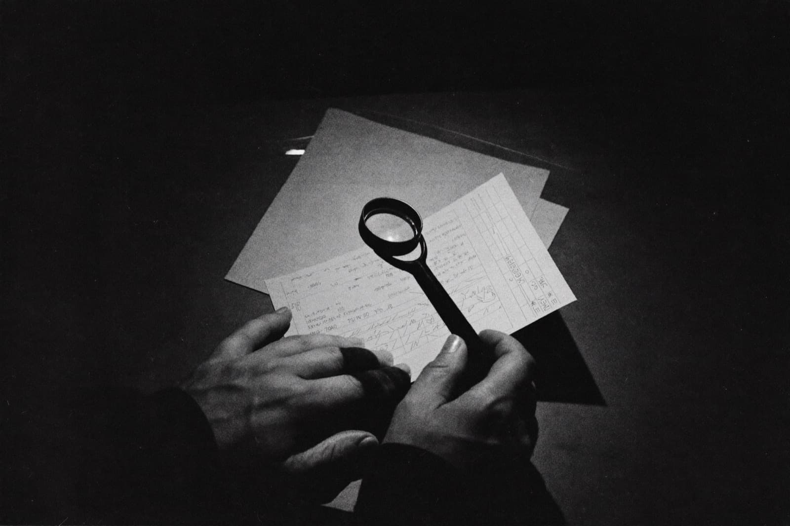 Hands examining a stack of receipts and printed charts through a loupe on a dark desk