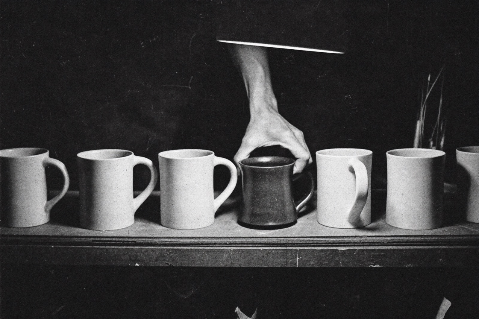 A hand pulling the one hand-thrown dark mug forward from a shelf of identical white mugs